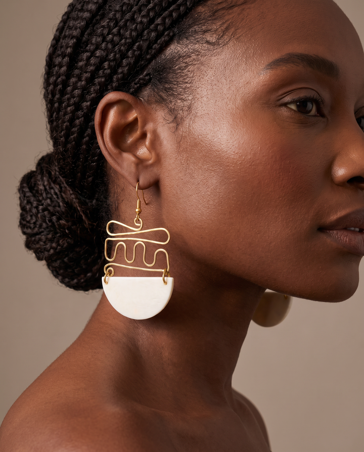 A close-up side profile of a woman wearing a large geometric dangle earring. The earring features a gold-tone wire design with a wavy pattern and a white semi-circular disc at the bottom, set against a warm beige background.